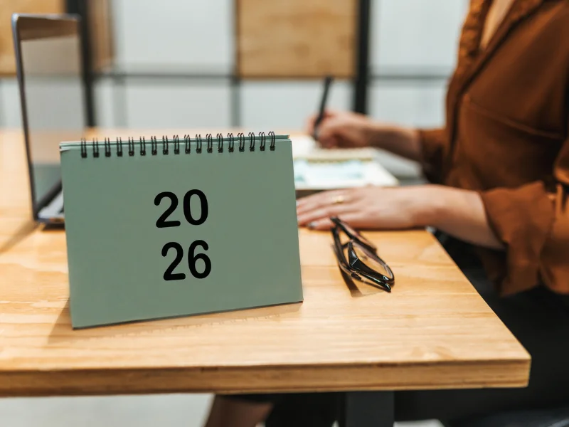 Visão detalhada de uma mesa de trabalho com laptop, calendário "2026" e uma mão escrevendo anotações em um caderno espiral com post-its.