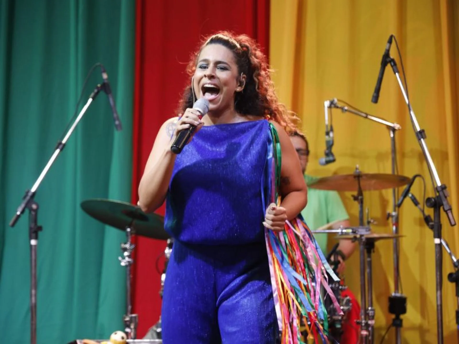 Uma mulher em um macacão azul canta no palco, segurando um microfone e fitas coloridas. Atrás dela, há uma bateria e cortinas verdes, vermelhas e amarelas.