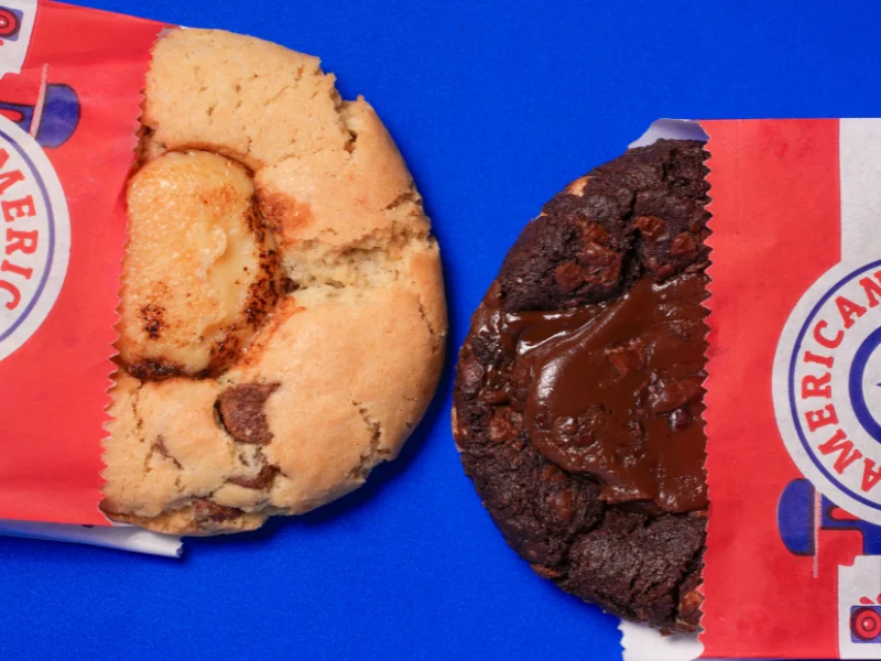 Dois cookies grandes saindo de embalagens vermelhas da American Cookies sobre um fundo azul; um é de massa clara com gotas e o outro é de chocolate com recheio fluido.