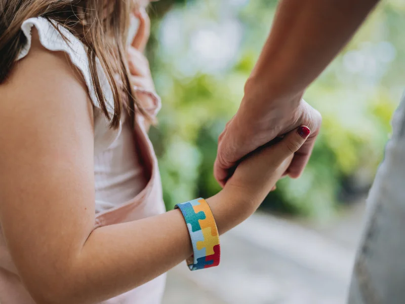 A imagem mostra o detalhe de uma criança usando a pulseira da conscientização do Transtorno do Espectro Autista (TEA) enquanto segura a mão de um adulto.