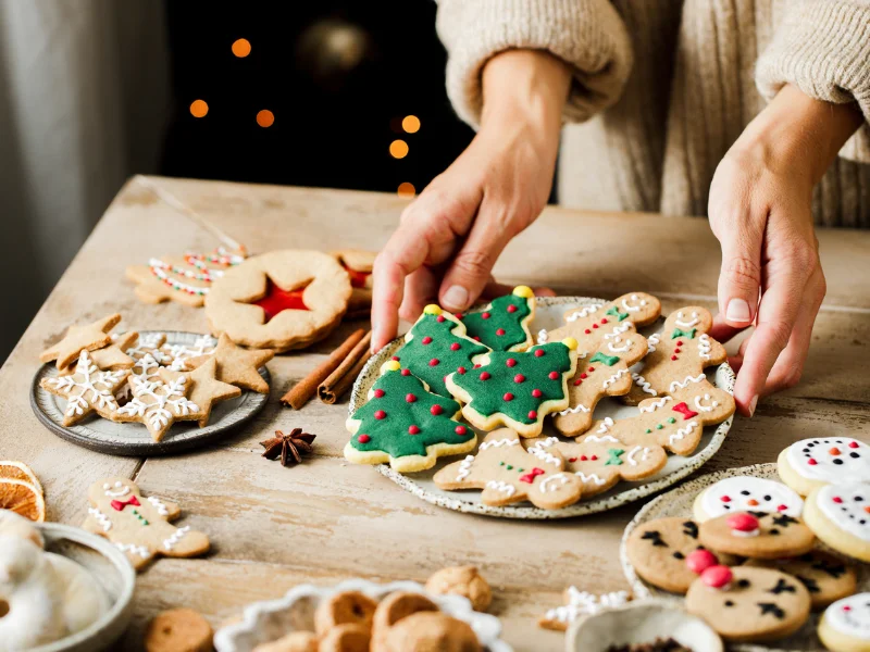Uma pessoa segura um prato cheio de biscoitos de gengibre decorados em formato de árvore de Natal e bonecos, sobre uma mesa de madeira rústica com outros biscoitos e especiarias.