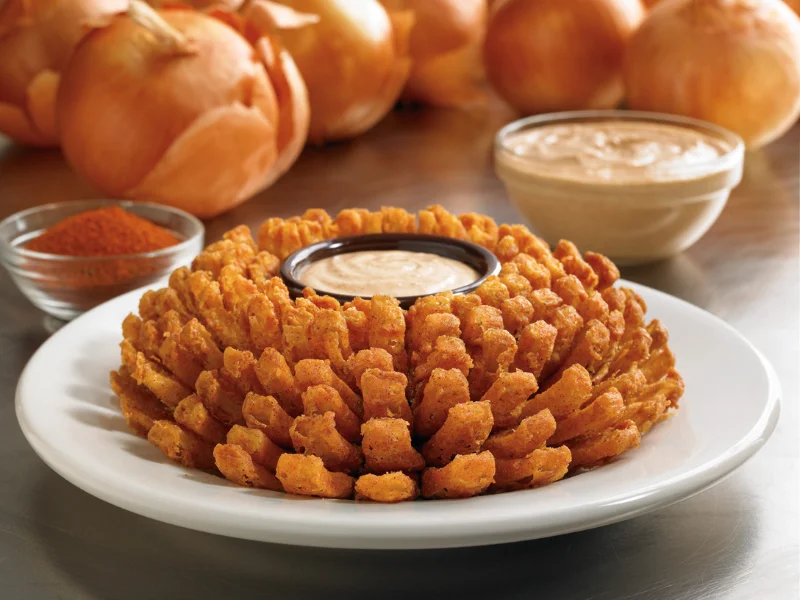 Fotografia em close de uma Bloomin' Onion, cebola empanada e frita em formato de flor, servida com molho central em um prato branco. Ao fundo, veem-se cebolas cruas e temperos.