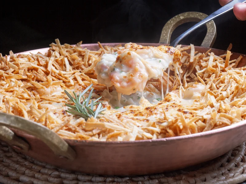Camarões gratinados com molho cremoso em uma panela de cobre, rodeados por uma borda crocante de batata palha, sendo servido com uma colher.