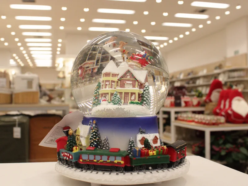 Um globo de neve grande com uma cena de casas nevadas e árvores, montado em uma base circular decorada com um trem de Natal colorido.