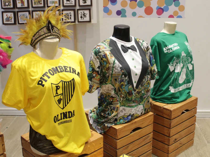 Três manequins exibindo as camisetas dos blocos Pitombeira (amarela), Homem da Meia-Noite (estilo fraque) e Amantes de Glória (verde).