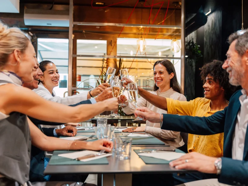 Seis pessoas de diversas idades e etnias estão sentadas em uma mesa de restaurante, sorrindo e fazendo um brinde com taças de vinho espumante. O ambiente é moderno e bem iluminado.