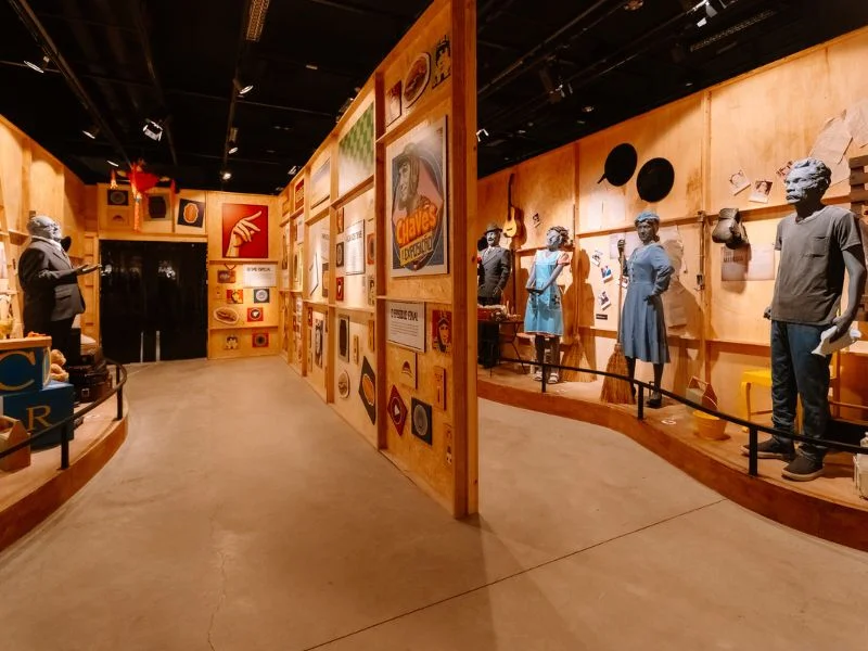 Fotografia colorida de uma exposição temática sobre o seriado "Chaves". O ambiente possui paredes de madeira clara repletas de quadros e cartazes, além de estátuas cinzas em tamanho real de personagens como Dona Florinda, Bruxa do 71 e Seu Madruga.