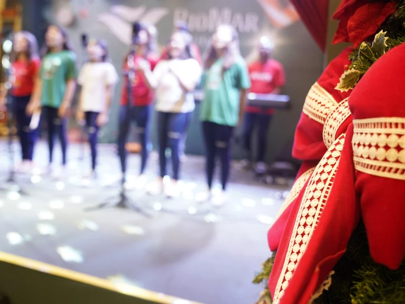Um grupo de jovens, em foco suave, cantando em um palco iluminado vestindo camisetas coloridas de Natal. Um grande laço vermelho e branco, parte da decoração, está nítido em primeiro plano à direita.