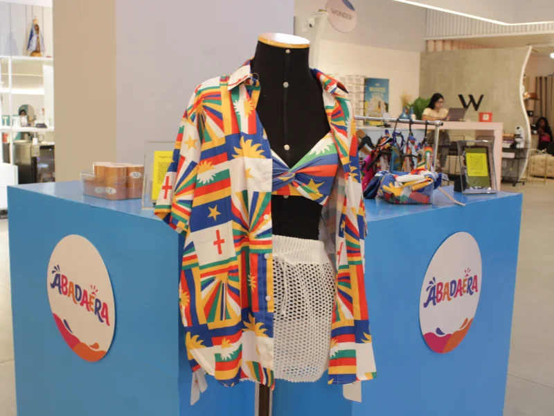 Manequim em um quiosque "Abadaêra" veste camisa aberta e top com estampa da bandeira pernambucana, combinados com uma saia branca de tela.