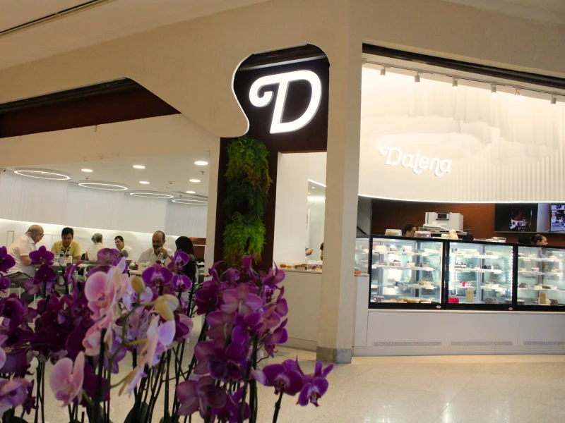 Entrada da cafeteria com um grande arranjo de orquídeas roxas em primeiro plano. A fachada apresenta a letra "D" estilizada, uma parede verde vertical e clientes sentados no interior.
