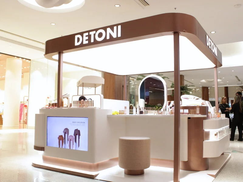 Vista geral de um quiosque de maquiagem da marca Detoni. O quiosque tem formato arredondado nas cores branco e cobre, com o nome "DETONI" em destaque e uma tela exibindo produtos.