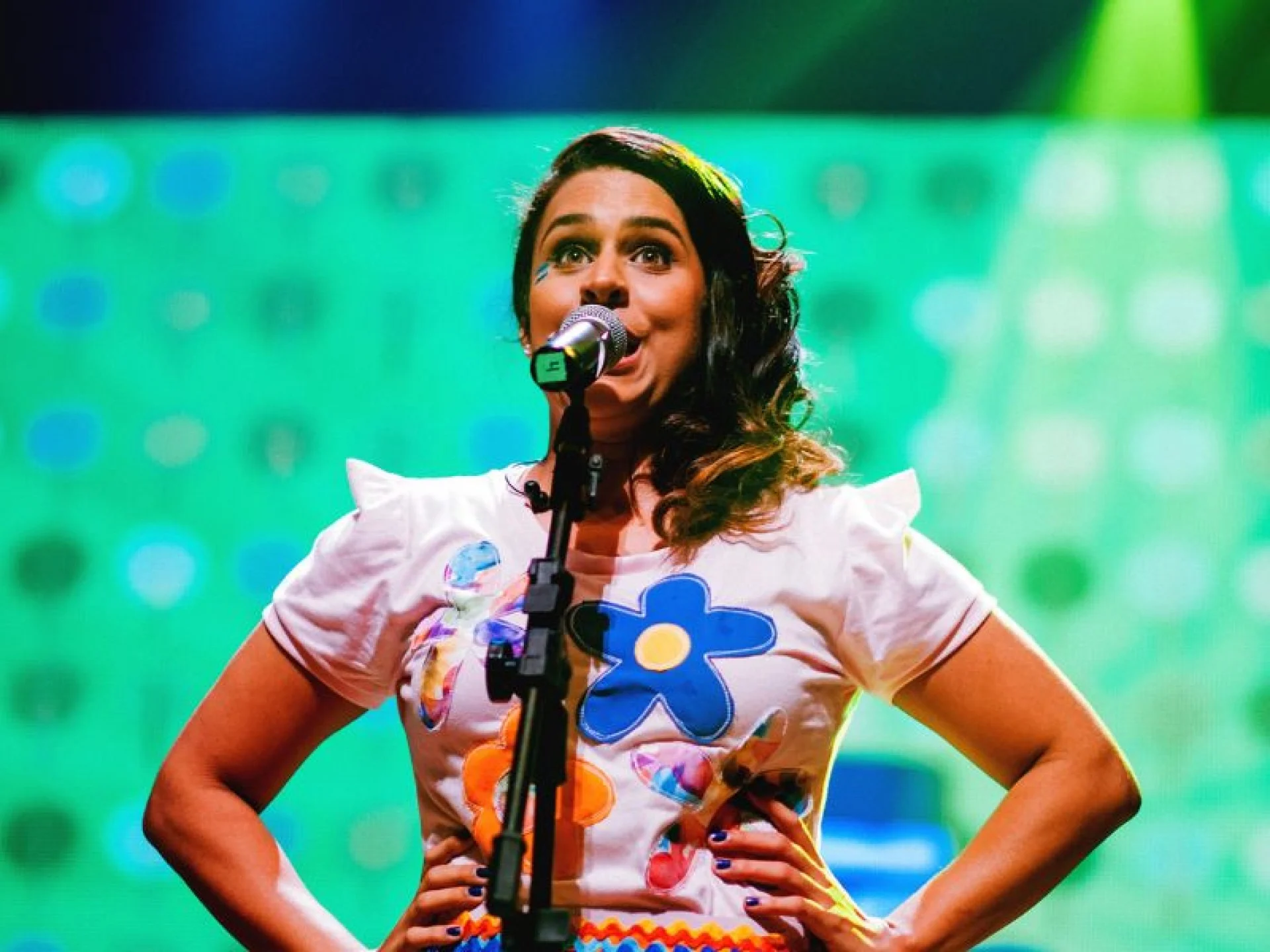 Uma mulher está no palco com as mãos nos quadris, falando ou cantando em um microfone. Ela veste uma camiseta branca com desenhos de flores coloridas, e o fundo é iluminado com luzes de palco verdes e amarelas.