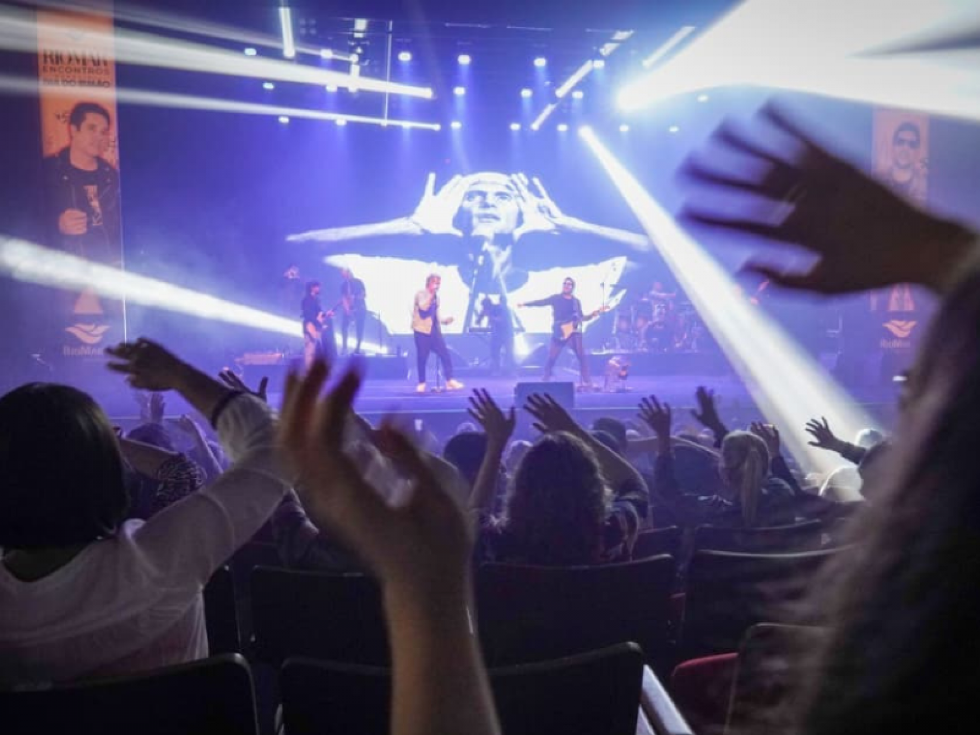 Plateia com mãos levantadas com o palco ao fundo.