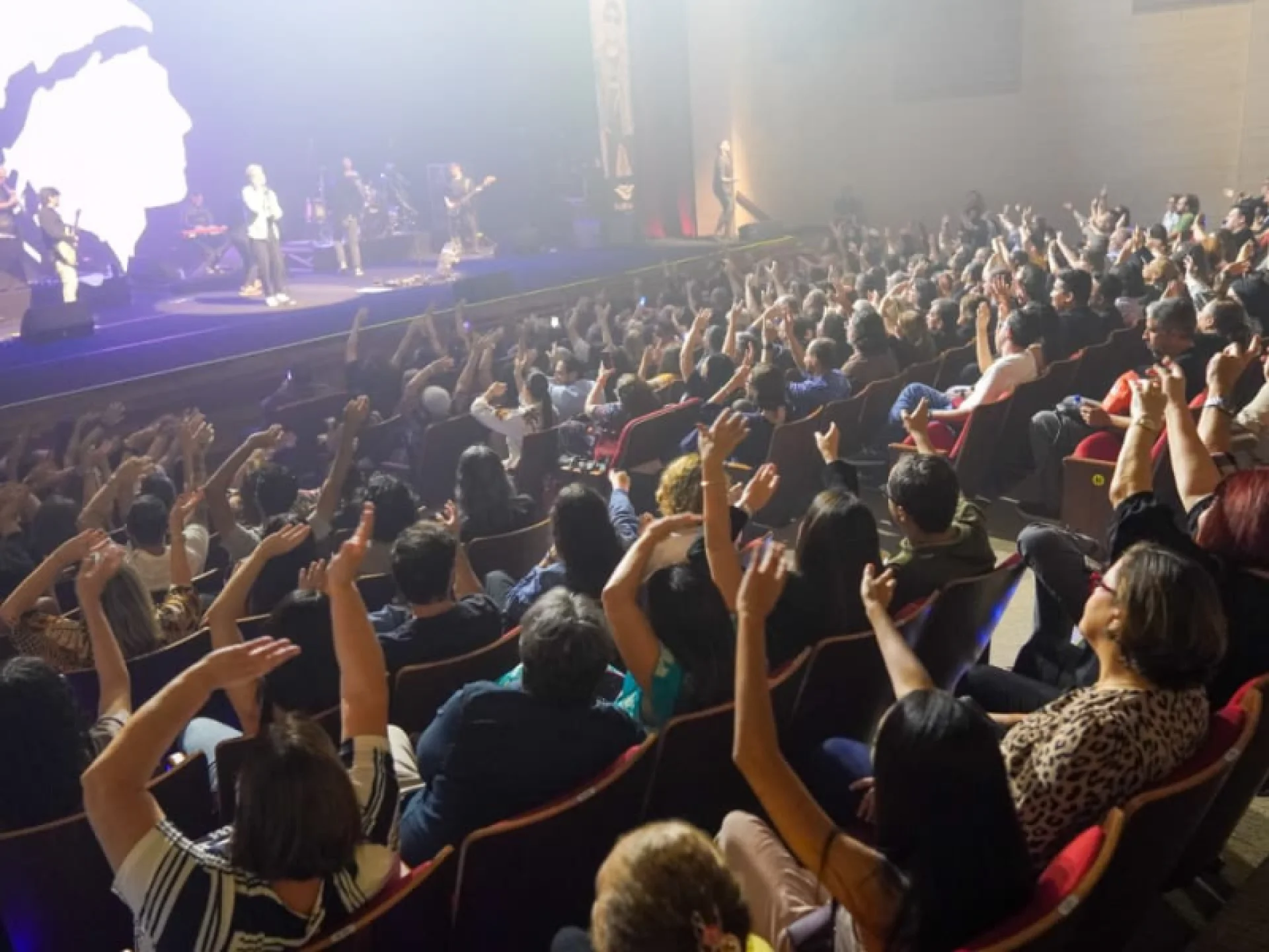 O público sentado no teatro com palco ao fundo.