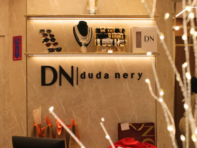 Detalhe da logomarca "DN | duda nery" em parede de mármore com prateleiras de acessórios iluminadas.