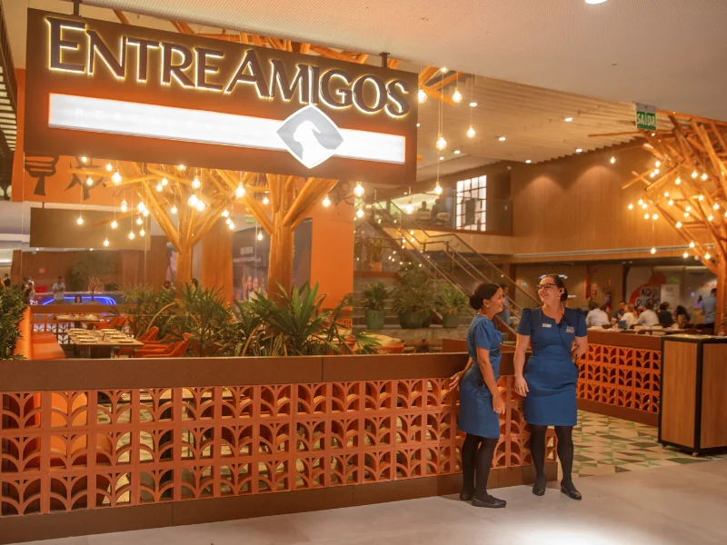 Entrada do restaurante com letreiro iluminado, decoração em tons terrosos e duas recepcionistas de uniforme azul sorrindo na recepção.