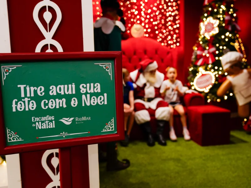 Close-up de uma placa verde e vermelha que diz "Tire aqui sua foto com o Noel", com a cena da foto (Papai Noel sentado com crianças) desfocada ao fundo.
