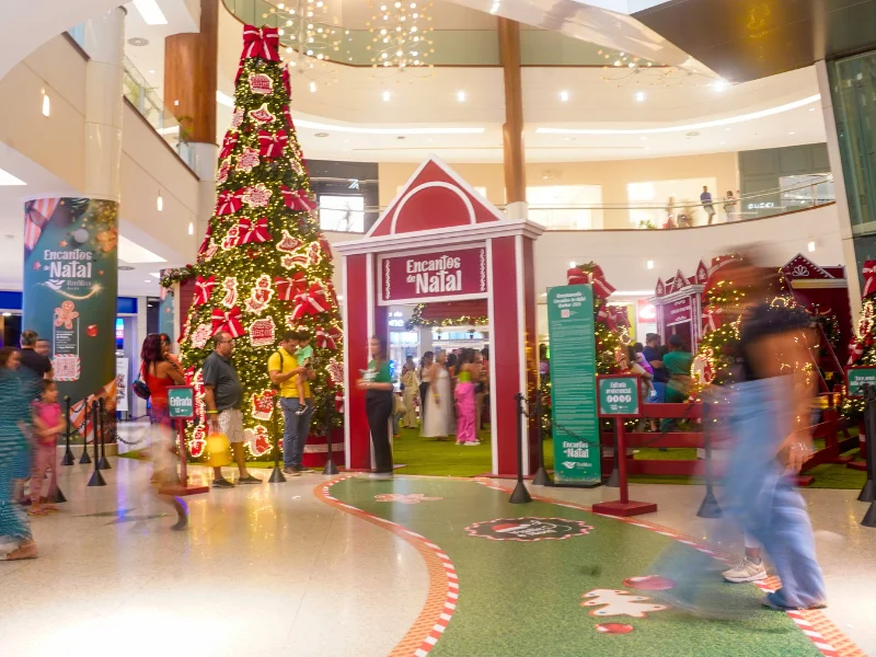 Tomada ampla e alta do espaço de eventos do shopping, destacando a área temática com piso de grama sintética, pequenas casinhas e uma grande placa de "Encantos de Natal" na parede de vidro.