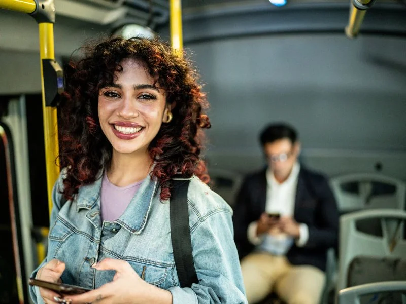 Mulher jovem com cabelos cacheados sorri enquanto segura um celular dentro de um ônibus. Ela usa jaqueta jeans e está próxima a um corrimão amarelo.