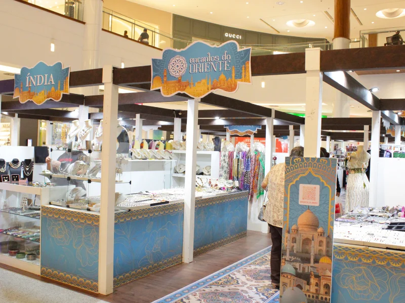 Estande da feira "Encantos do Oriente" em um shopping, com estrutura de madeira, balcões azuis decorados e exposição de joias e acessórios.
