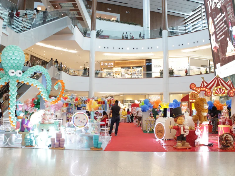 Vista ampla de um shopping com stands de festas infantis, destacando um polvo gigante de balões azuis à esquerda e um portal de circo à direita.