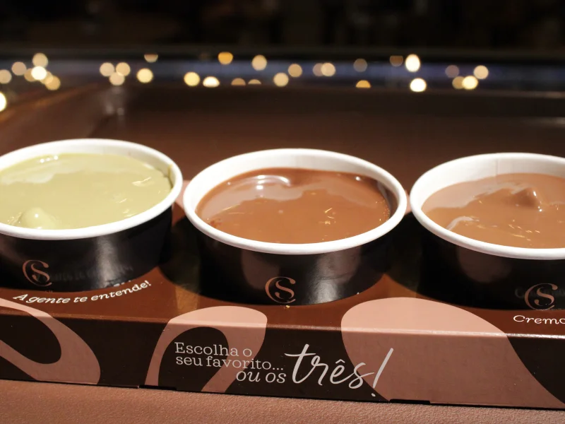 Três potes individuais de fondue, um pistache e dois de chocolate, estão dispostos lado a lado sobre uma embalagem promocional. A embalagem tem o texto "Escolha o seu favorito... ou os três!".
