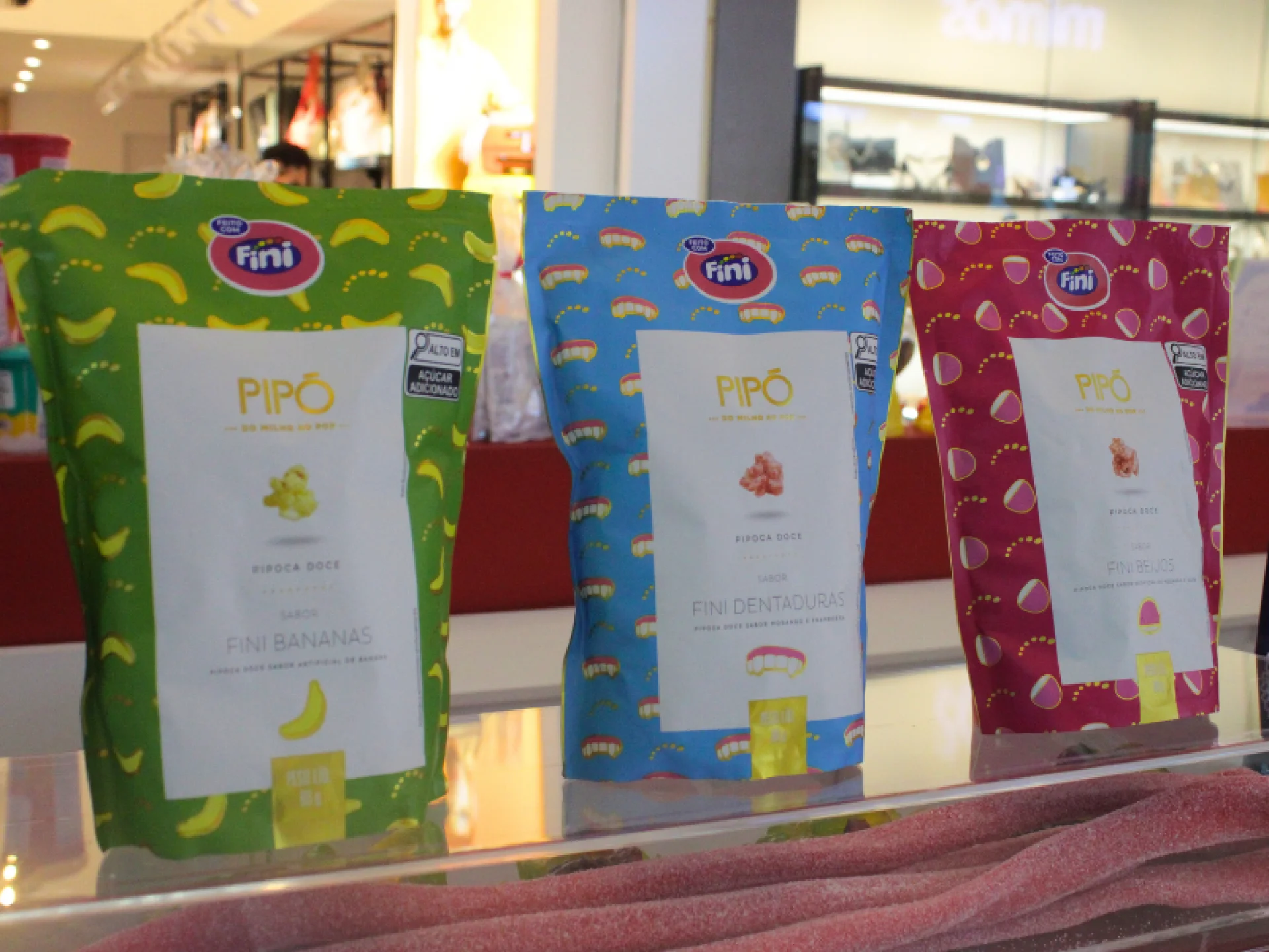 Três pacotes de pipoca Pipó Fini estão lado a lado em um balcão: bananas (verde), dentaduras (azul) e beijos (rosa).