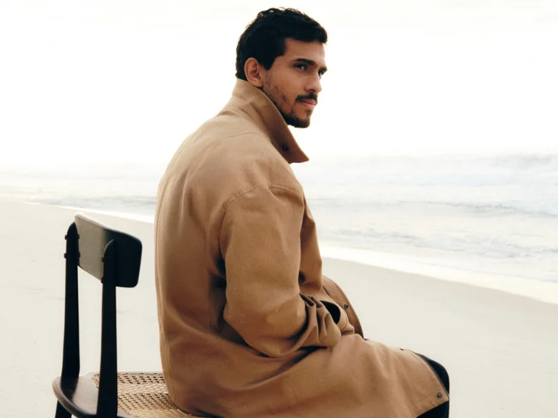Homem sentado de costas em uma cadeira na praia, olhando sobre o ombro; ele veste um casaco longo de cor caramelo.