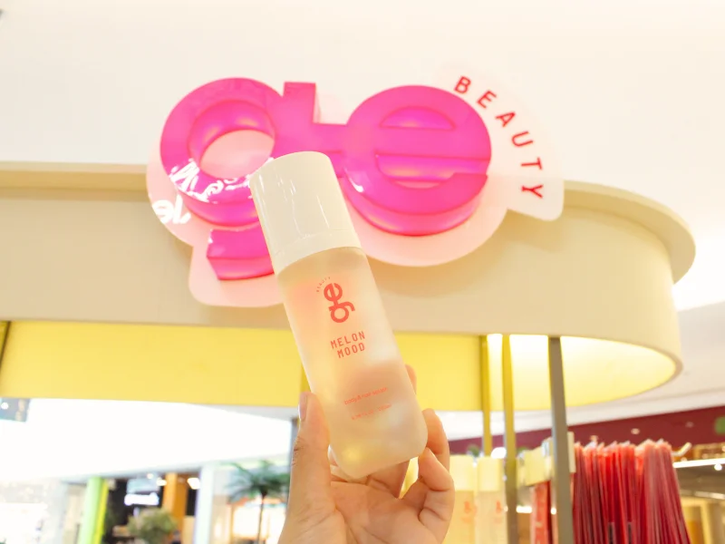A imagem mostra uma mão segurando um frasco de Body e Hair Splash com a logo da GE Beauty ao fundo em tom de rosa
