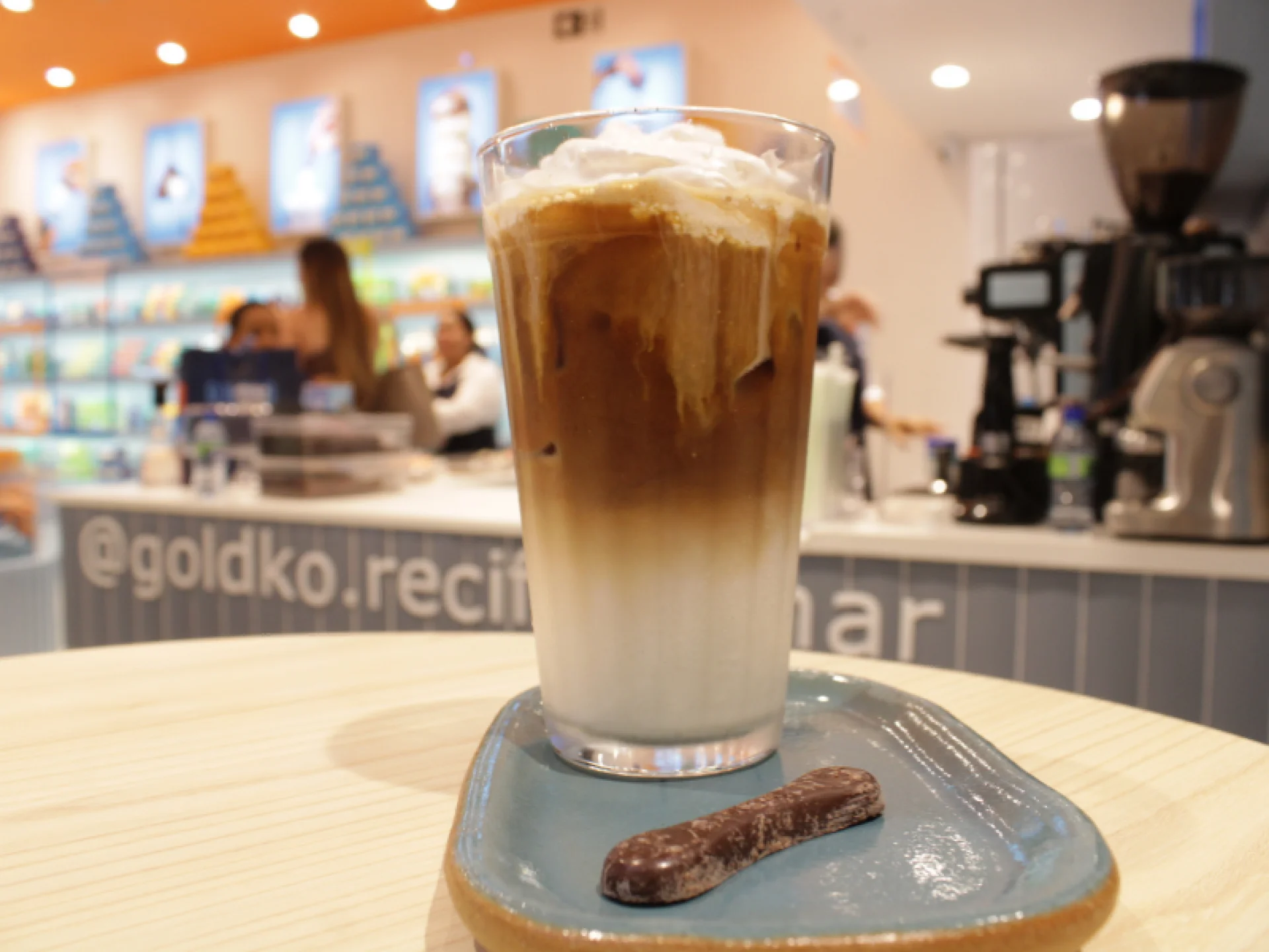 Copo com bebida gelada com café em um prato com um chocolate ao lado. O prato está em cima de uma mesa que tem a loja GoldKo de fundo.