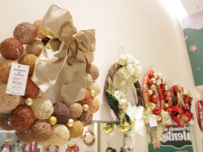 Em destaque, uma guirlanda feita de bolas de linha em tons terrosos com um laço dourado, exibida ao lado de outras guirlandas tradicionais em vermelho, dourado e verde, penduradas em uma parede branca.