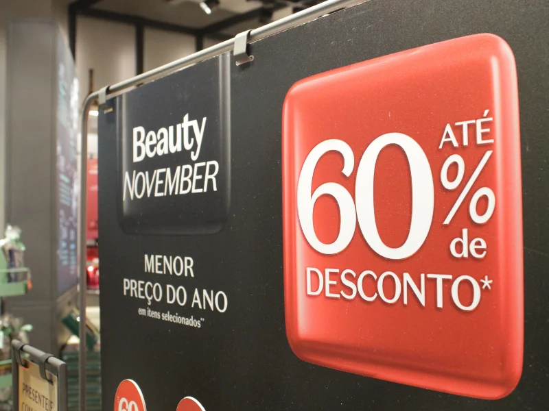 Um cartaz de promoção em uma loja anuncia o evento "Beauty November", com o "Menor Preço do Ano" em itens selecionados. Um grande quadrado vermelho ao lado destaca o desconto de "Até 60% de Desconto".