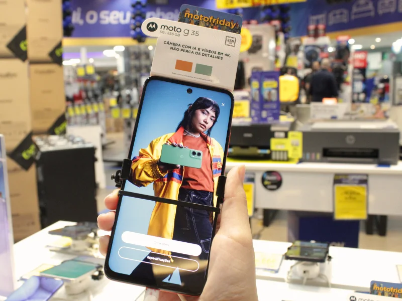 Uma mão segura um smartphone Motorola em uma loja, exibindo na tela uma modelo segurando outro celular. Um cartaz branco no topo indica ser um "moto g35" com câmera 4K, durante o evento "moto friday".