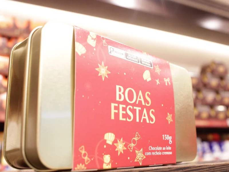 Lata metálica dourada com uma faixa vermelha onde se lê "BOAS FESTAS". Contém 150g de chocolate ao leite com recheio cremoso.