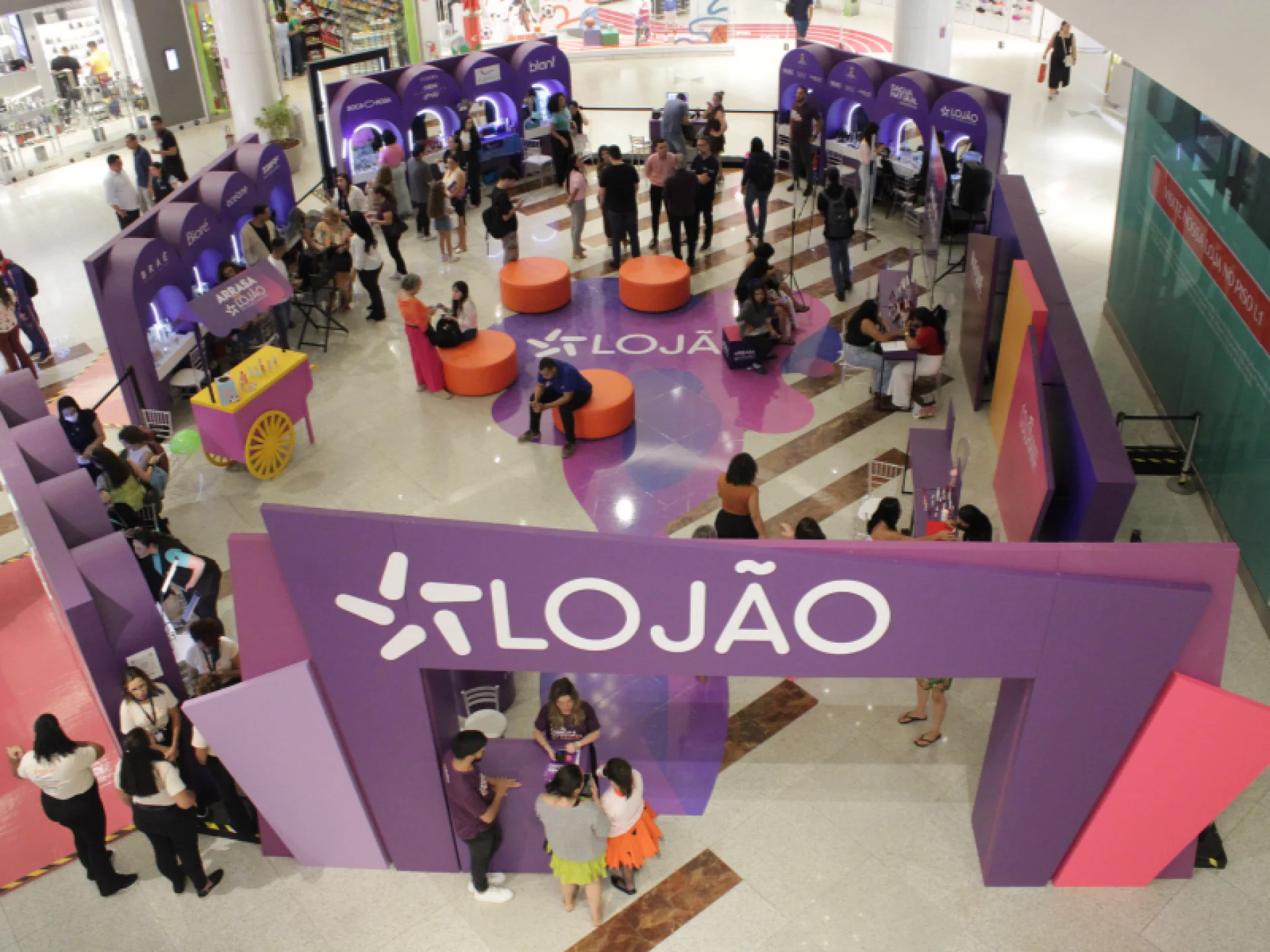 A imagem mostra uma visão aérea do estande "LOJÃO" em um shopping, com diversas estações de beleza roxas e rosas, cheias de público.