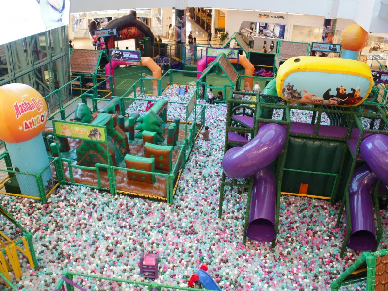 Vista aérea ampla do parque mostrando uma enorme piscina de bolinhas coloridas com escorregadores roxos e obstáculos infláveis verdes.