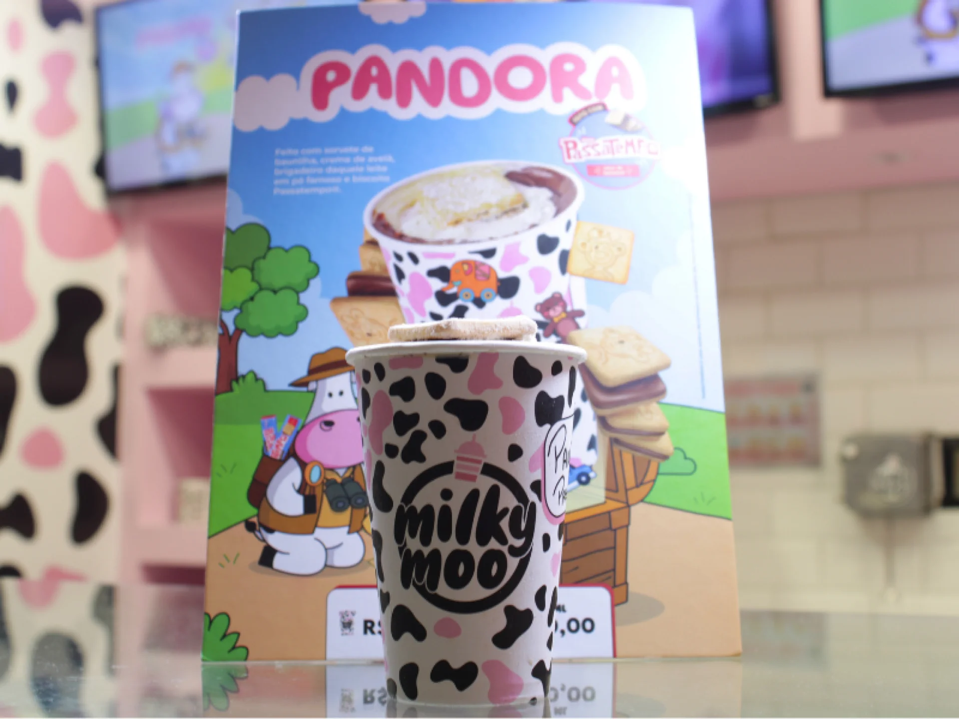 Foto de um copo de milkshake da marca Milky Moo sobre um balcão, com um grande cartaz de fundo que promove o sabor "Pandora Passatempo". Foto de um copo de milkshake da marca Milky Moo sobre um balcão, com um grande cartaz de fundo que promove o sabor "Pandora Passatempo".