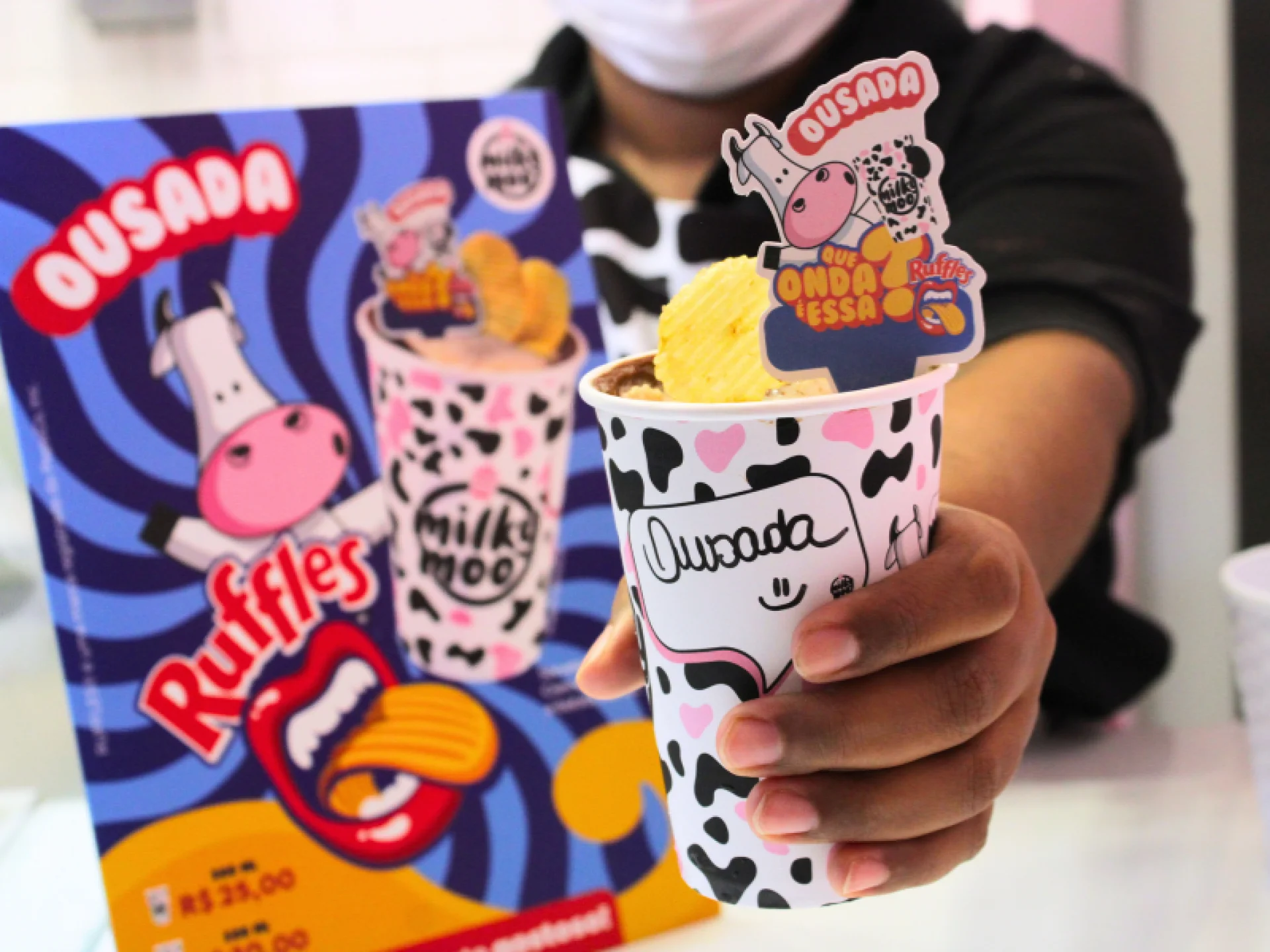 Copo de de milkshake segurado em primeiro plano com um cartaz do sabor 'Ousada', que é o milkshake de Ruffles com Nutella, ao fundo.