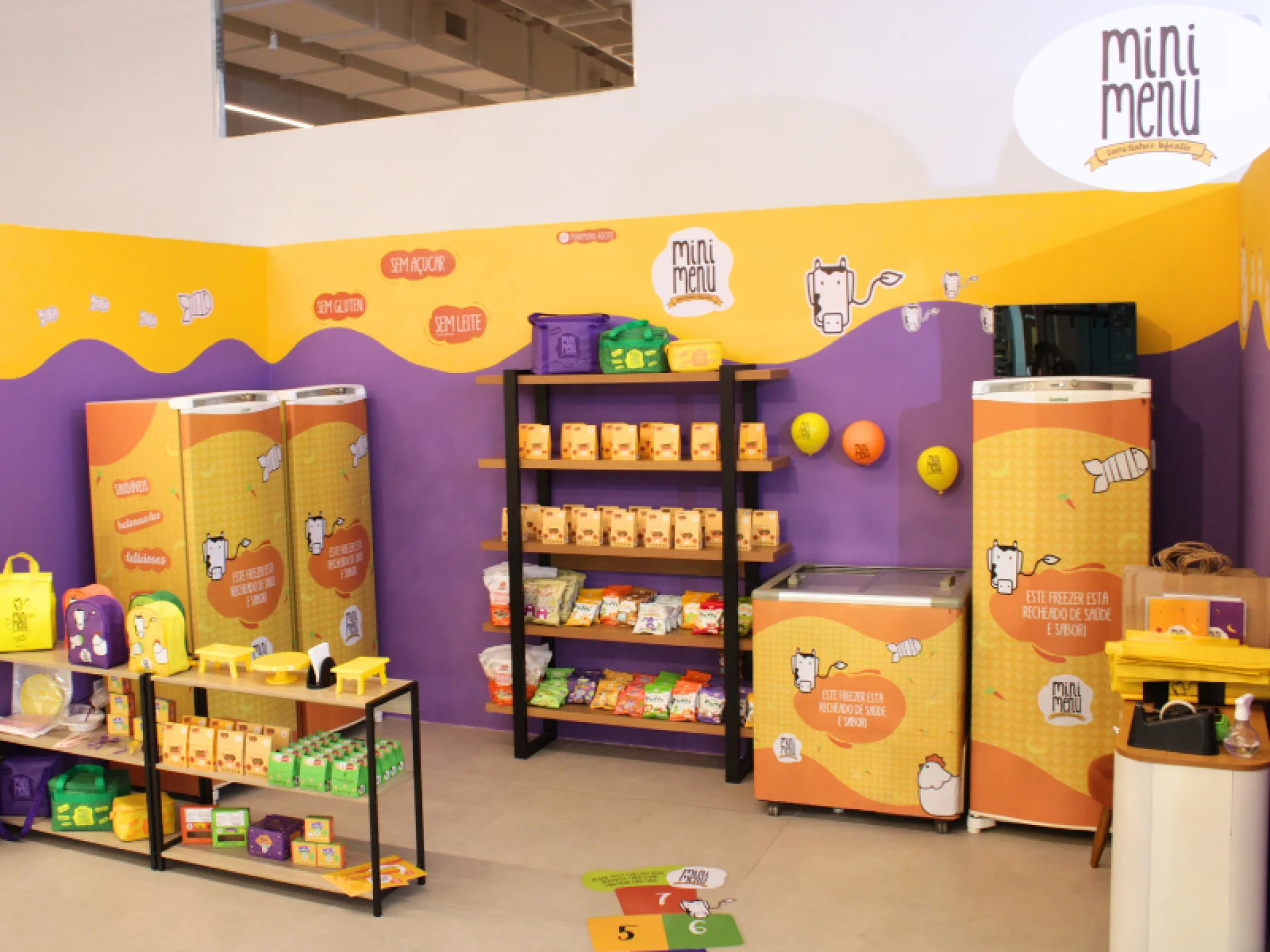 Um canto de loja com cores vivas, com paredes amarelas e roxas, exibindo a marca "Mini Menu". As prateleiras contêm lanches, coolers, mochilas e produtos com personagens de desenhos animados e designs divertidos.