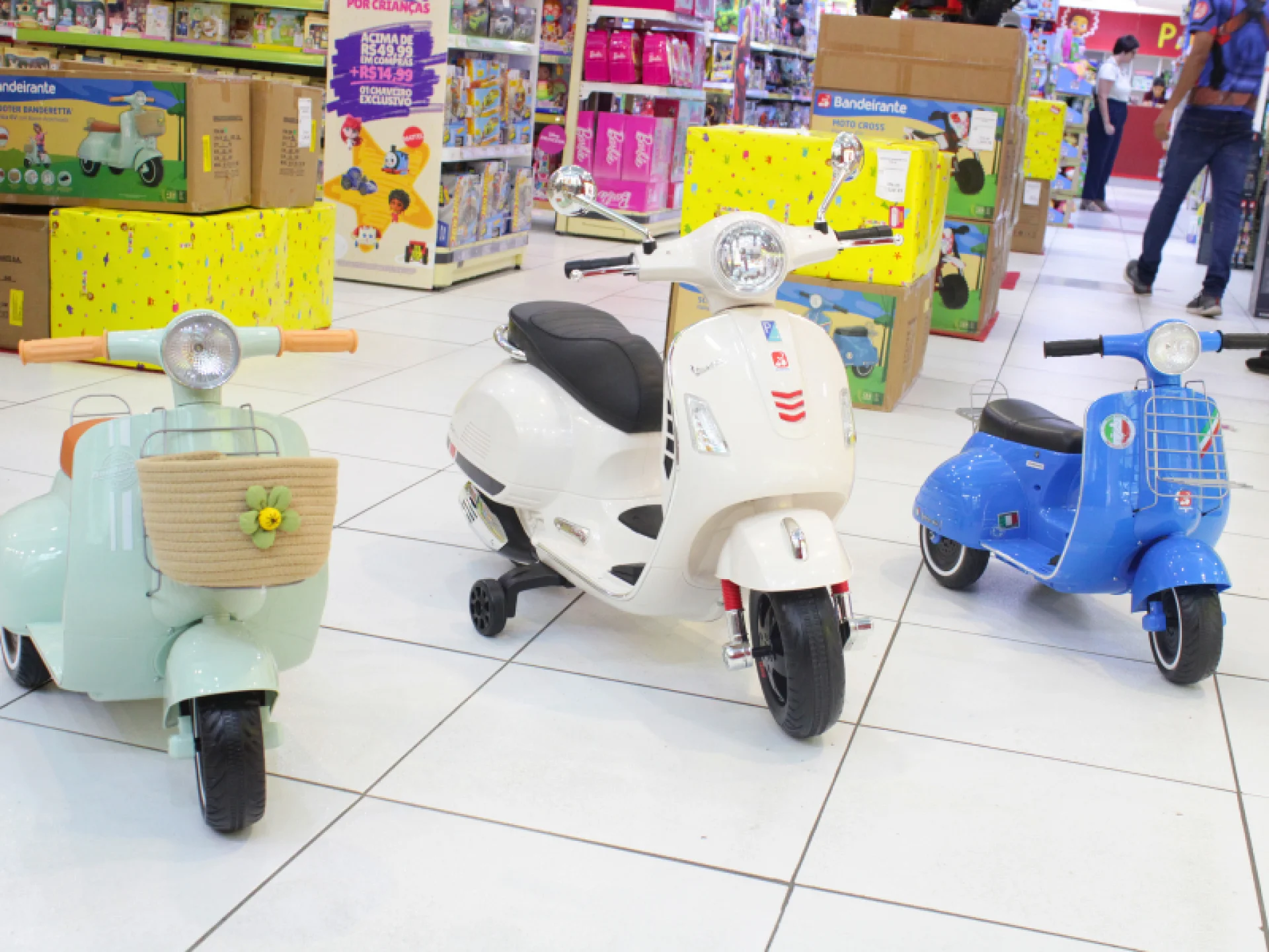 Três motos elétricas do tipo Vespa de brinquedo coloridos - verde-menta, branco e azul - estão expostos em um piso de azulejos brancos em uma loja de brinquedos.