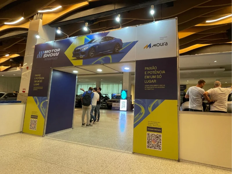Entrada do evento Motor Show Pernambuco com um arco azul e amarelo. O banner principal exibe um carro esportivo azul e a logomarca da patrocinadora Moura.