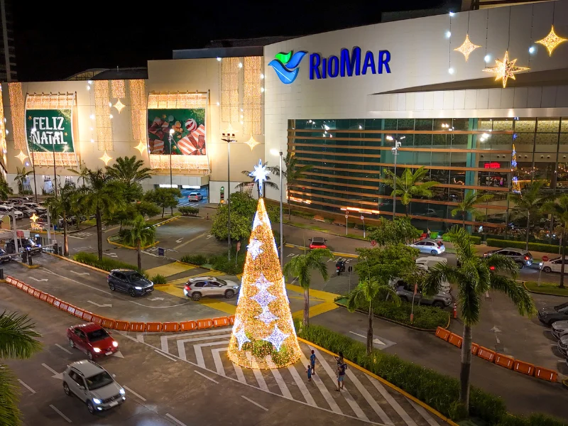 Vista noturna da fachada principal do RioMar Recife decorada para o Natal, com painéis de "Feliz Natal" e estrelas iluminadas. Uma grande árvore de Natal cônica e luminosa se destaca na via de acesso do shopping.
