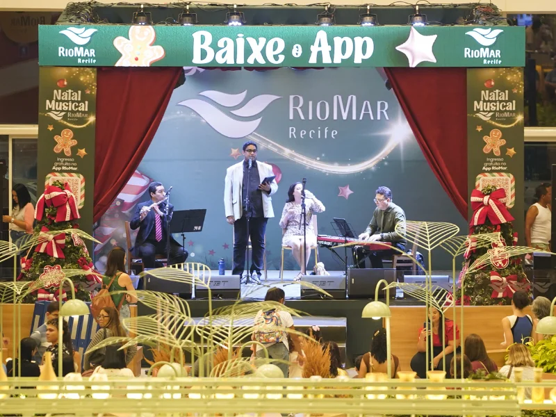 Palco do evento "Natal Musical RioMar Recife", com uma banda e cantores se apresentando sob cortinas vermelhas e um banner de Natal, com o público assistindo na frente.