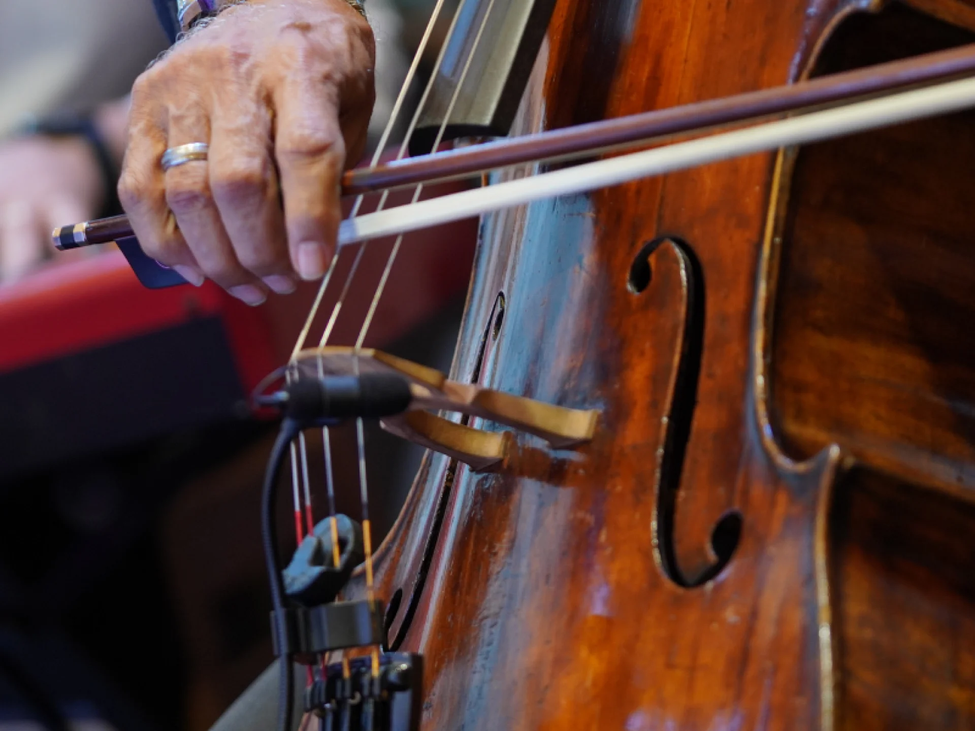 Close-up detalhado da m&atilde;o de um m&uacute;sico segurando o arco sobre as cordas de um violoncelo de madeira escura. &Eacute; poss&iacute;vel ver um anel de prata no dedo do m&uacute;sico.