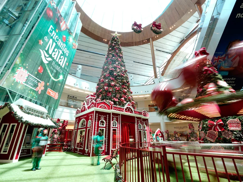 Ampla visão do átrio do shopping com a alta árvore de Natal central sobre uma base vermelha de biscoito, e decorações de casinhas menores ao redor. Uma faixa vermelha e verde com o tema "Encantos de Natal RioMar" está à esquerda.