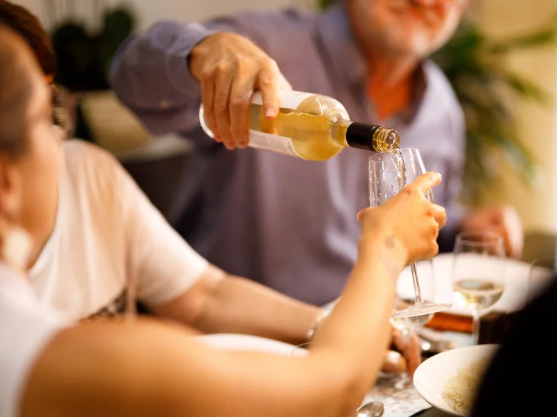 Em um jantar compartilhado, uma pessoa serve vinho branco em uma taça segurada por outra, criando uma atmosfera acolhedora.
