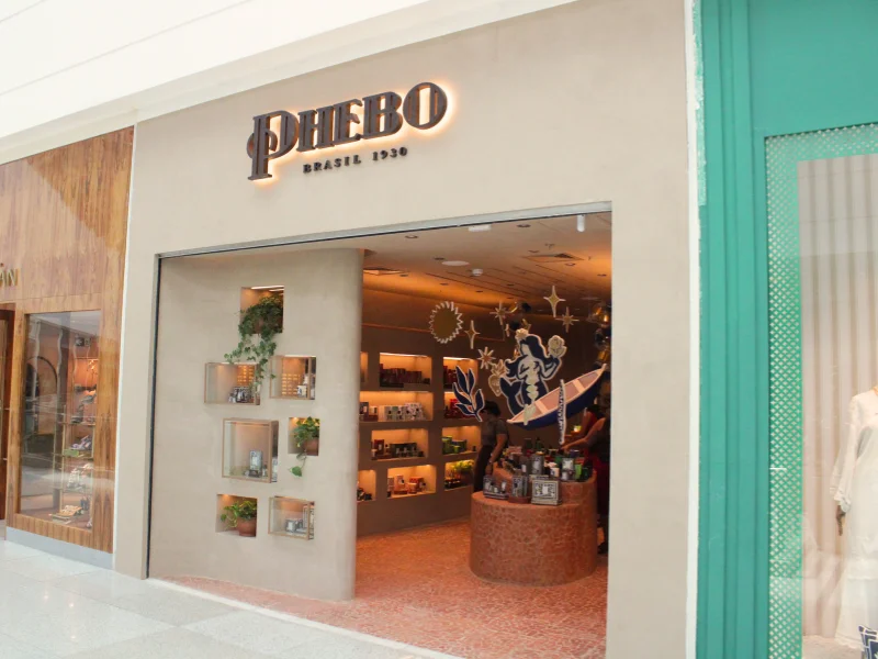 Fachada da loja Phebo, com o letreiro em relevo e nichos decorativos com plantas na entrada. O interior apresenta um balcão redondo e decoração temática iluminada.