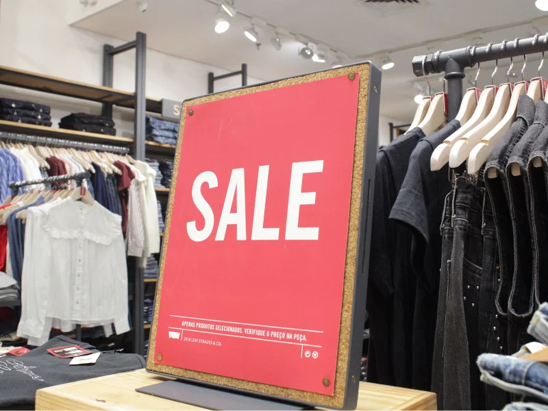 Placa vermelha com a palavra "SALE" em branco em destaque, indicando uma promoção em uma loja de roupas. A placa está posicionada em frente a uma arara com roupas escuras e outras peças desfocadas ao fundo.
