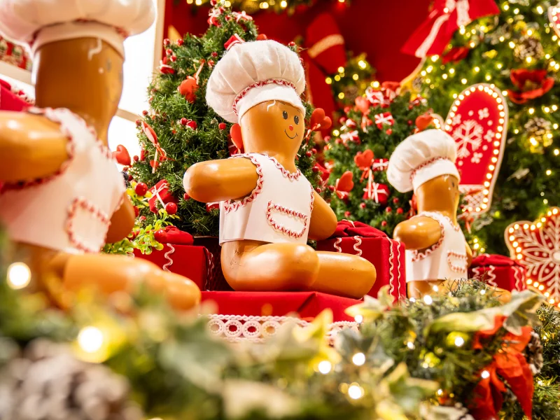 Cena de decoração natalina com grandes bonecos de Biscoito de Gengibre vestidos de chef, sentados em caixas vermelhas e cercados por árvores de Natal e luzes.
