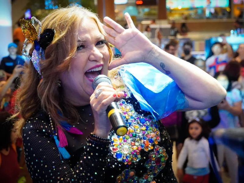 Na imagem, aparece a cantora Nena Queiroga, em primeiro plano, com expressão de pura alegria e entusiasmo com a plateia desfocada no fundo.